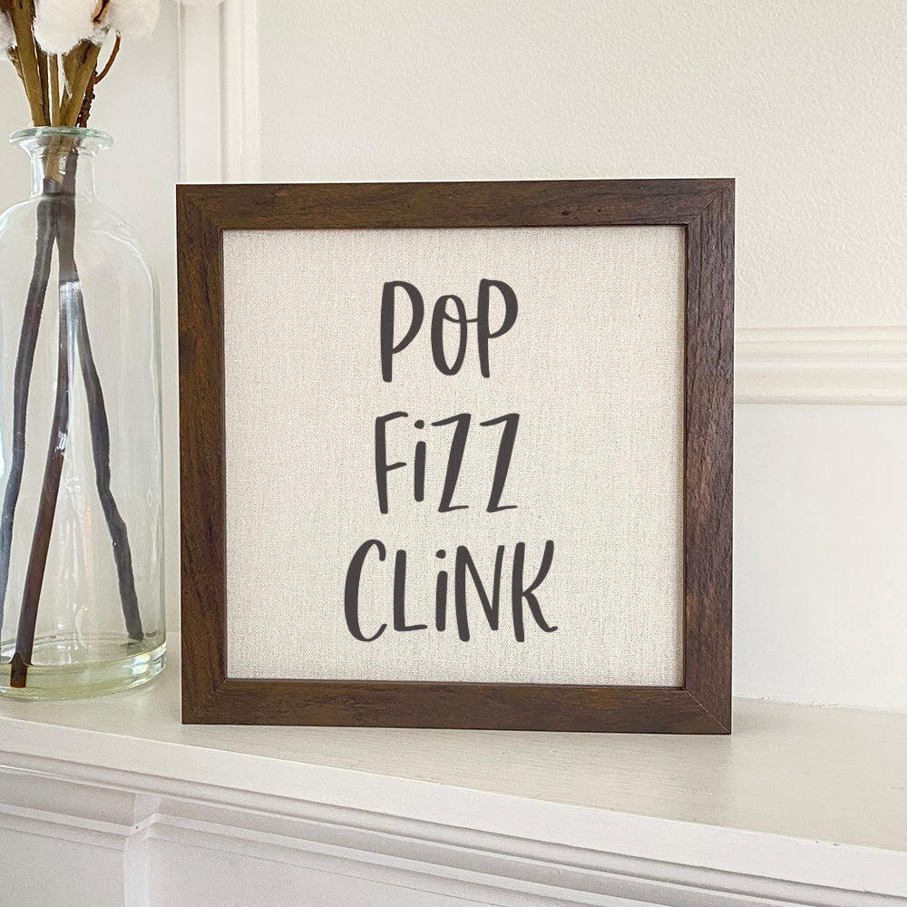 Pop Fizz Clink framed sign with a stylish wood frame, featuring a linen-look background and eco-friendly ink printing.