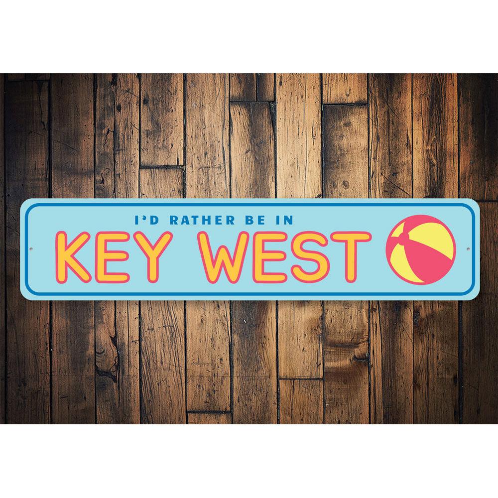 A decorative metal sign reading 'Rather Be in Key West', featuring vibrant colors and a beach-themed design, perfect for coastal decor.