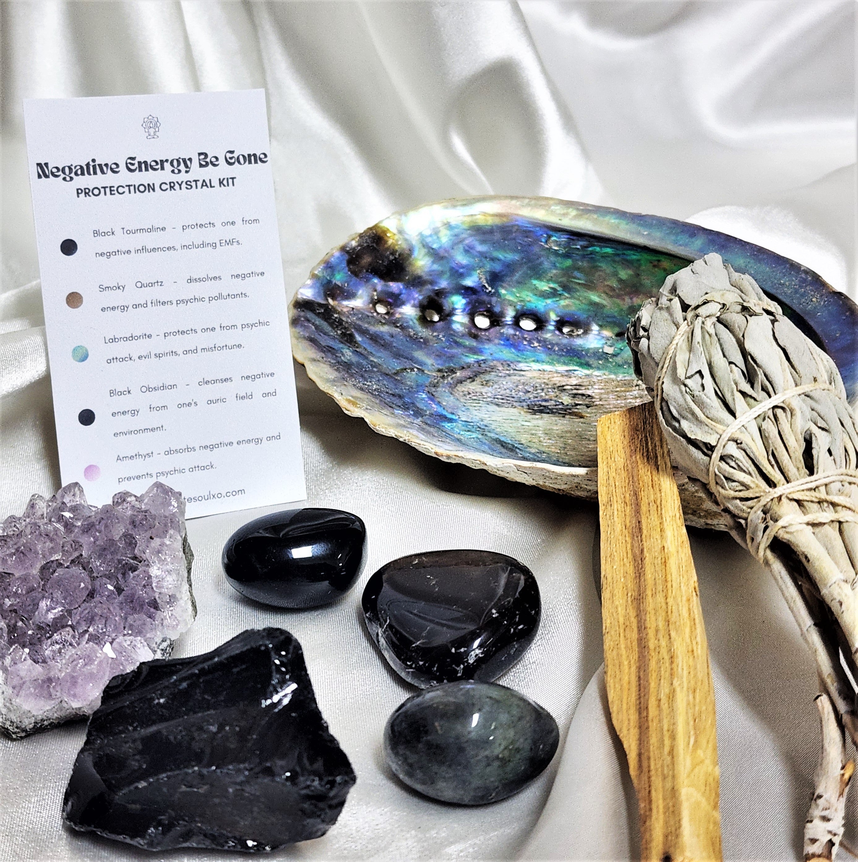 A beautifully arranged Protection Smudge Kit featuring a large sage smudge stick, palo santo wood, an abalone shell, and various protective crystals including smoky quartz and black tourmaline.