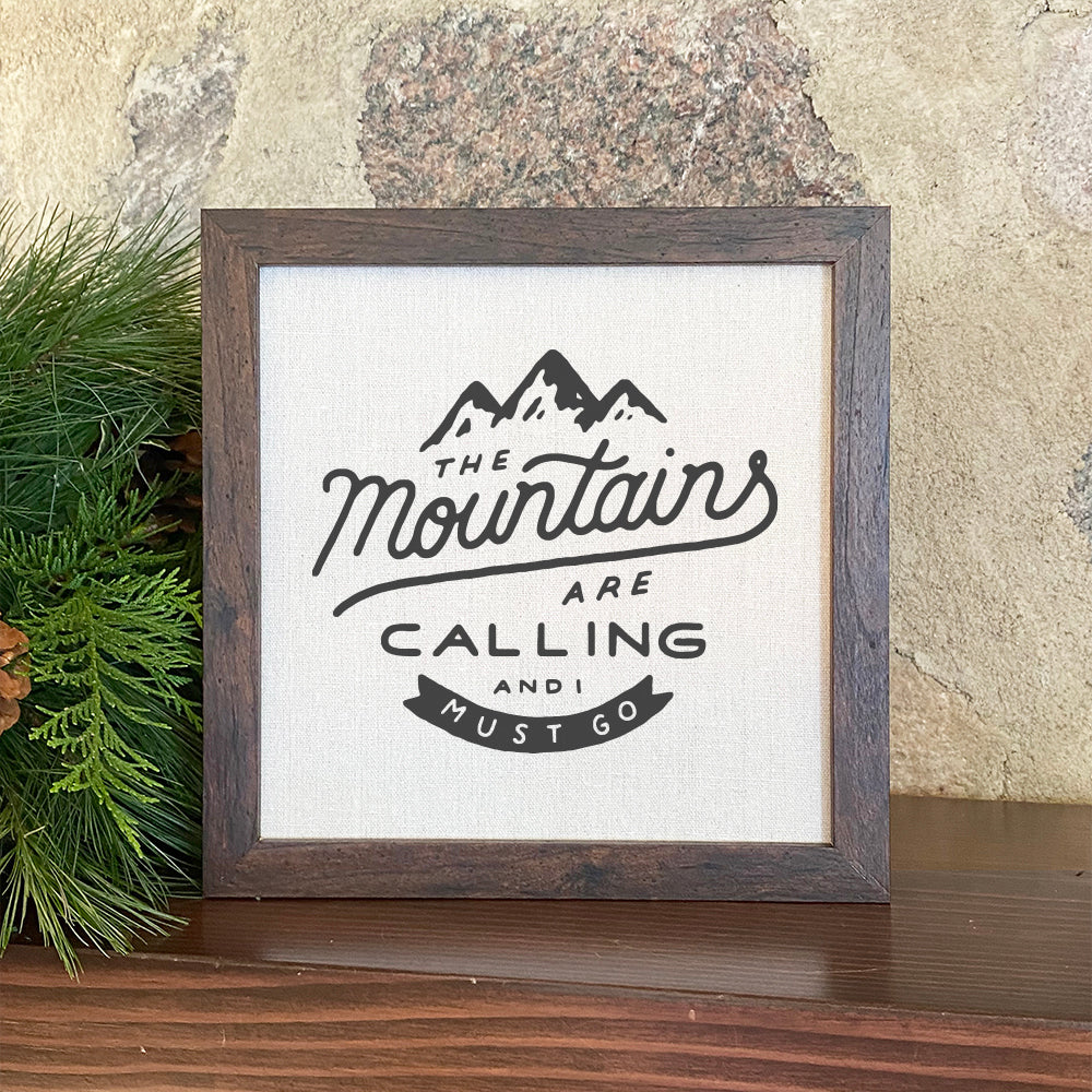 Framed sign featuring the phrase 'The Mountains are Calling' with a rustic wood frame and linen-look background.