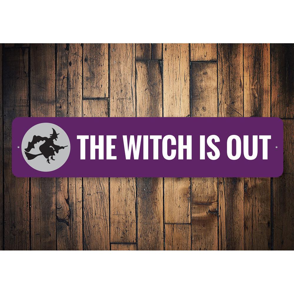 A decorative Halloween sign reading 'The Witch is OUT', made of high-quality aluminum, featuring spooky design elements.