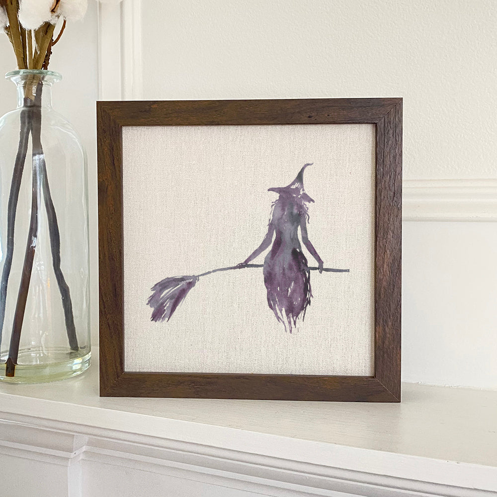 A framed sign featuring a witch silhouette, set in a stylish wood frame with a linen-look background.