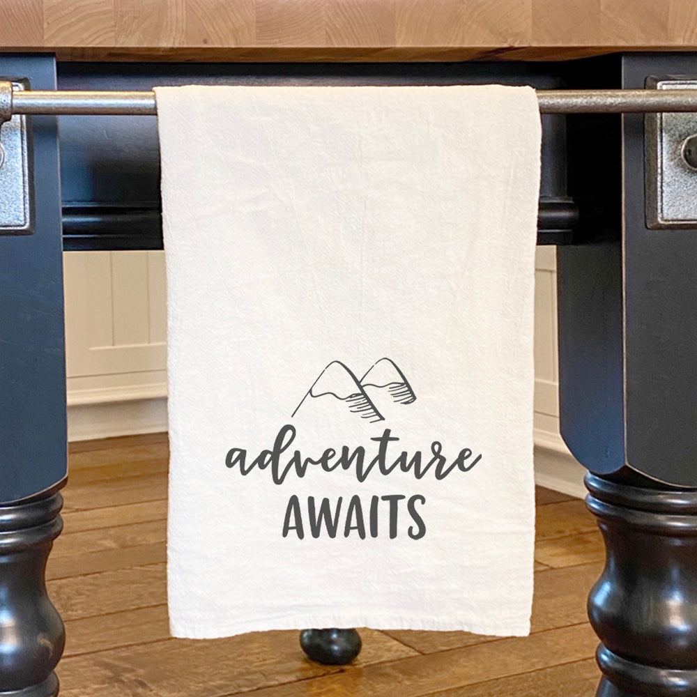 Adventure Awaits cotton tea towel featuring a vibrant mountain design, made from 100% absorbent cotton with hemmed edges.