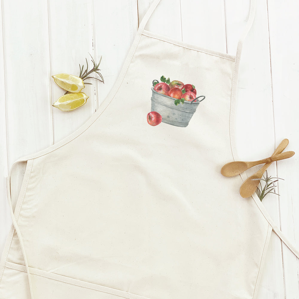 A stylish women's apron featuring a bucket of red apples design, made from durable cotton canvas with adjustable neck and twill ties.