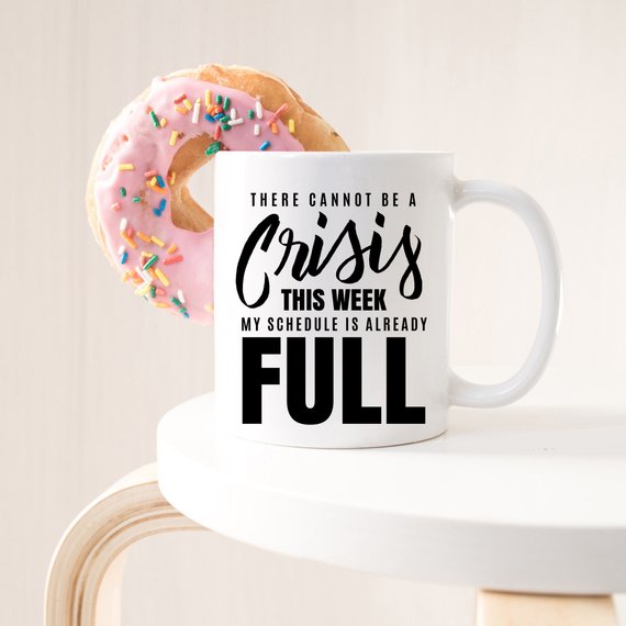 A humorous coffee mug with the phrase 'There Cannot Be A Crisis This Week' printed on it, showcasing a fun design perfect for office gifts.