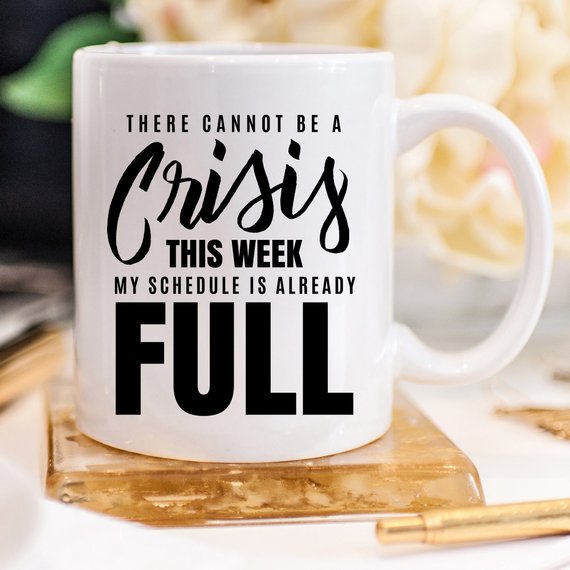 A humorous coffee mug with the phrase 'There Cannot Be A Crisis This Week' printed on it, showcasing a fun design perfect for office gifts.