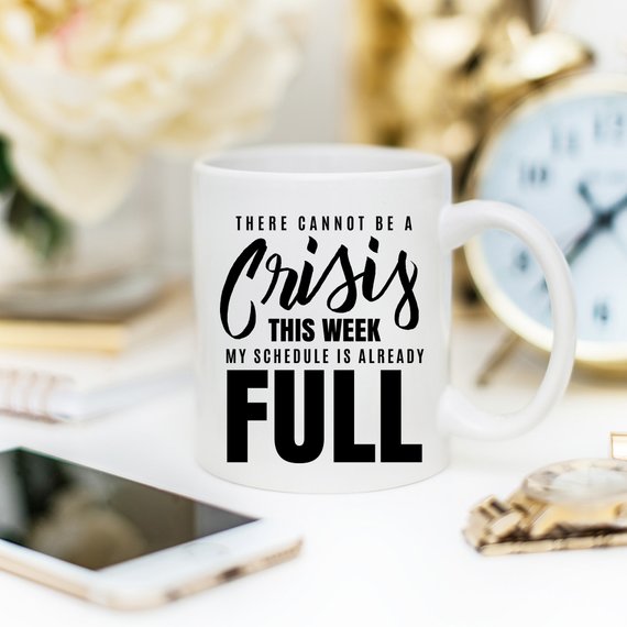 A humorous coffee mug with the phrase 'There Cannot Be A Crisis This Week' printed on it, showcasing a fun design perfect for office gifts.