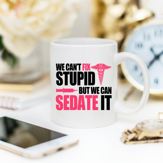 A humorous coffee mug designed for nurses, featuring the text 'We Can't Fix Stupid, But We Can Sedate It' in bold, readable font on a white ceramic background.