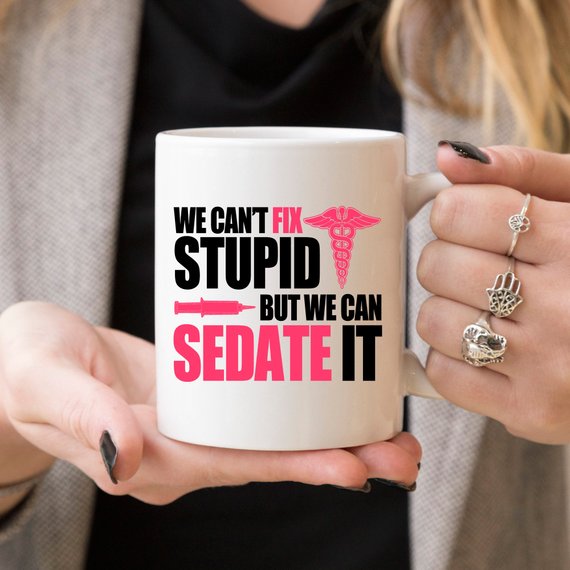 A humorous coffee mug designed for nurses, featuring the text 'We Can't Fix Stupid, But We Can Sedate It' in bold, readable font on a white ceramic background.