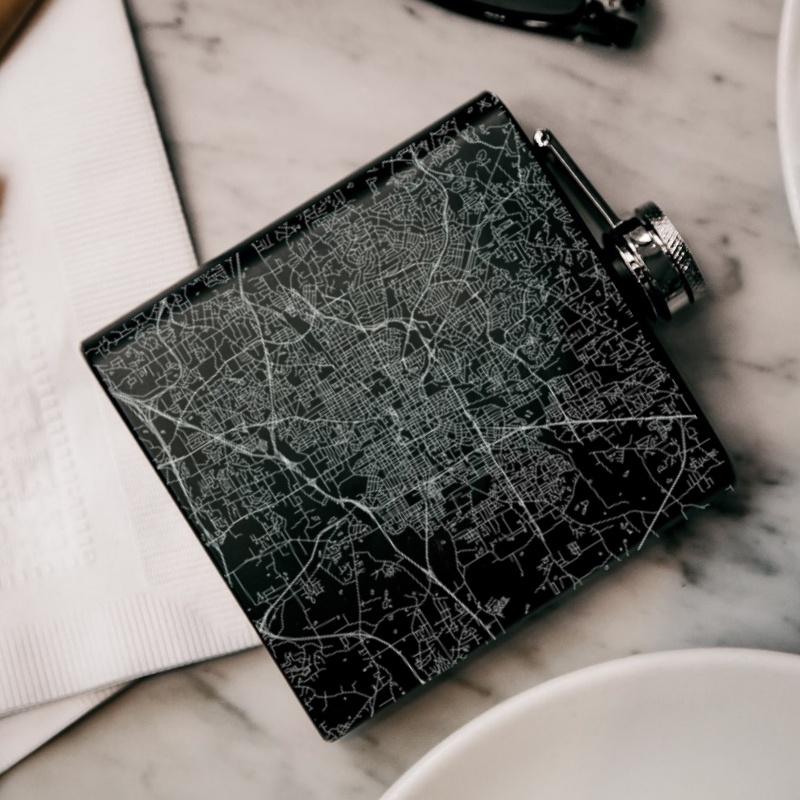 Matte black hip flask featuring a custom engraved map of Greensboro, North Carolina, with coordinates, alongside a canvas bag and funnel.