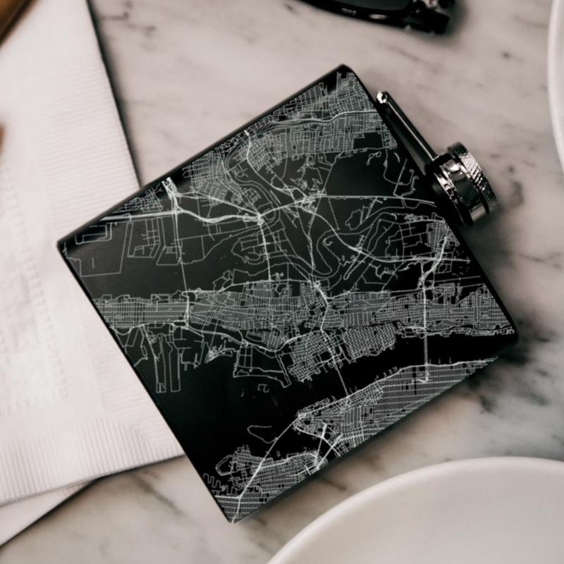 Matte black hip flask featuring a custom engraved map of Jersey City, New Jersey, with coordinates, alongside a canvas bag and funnel.