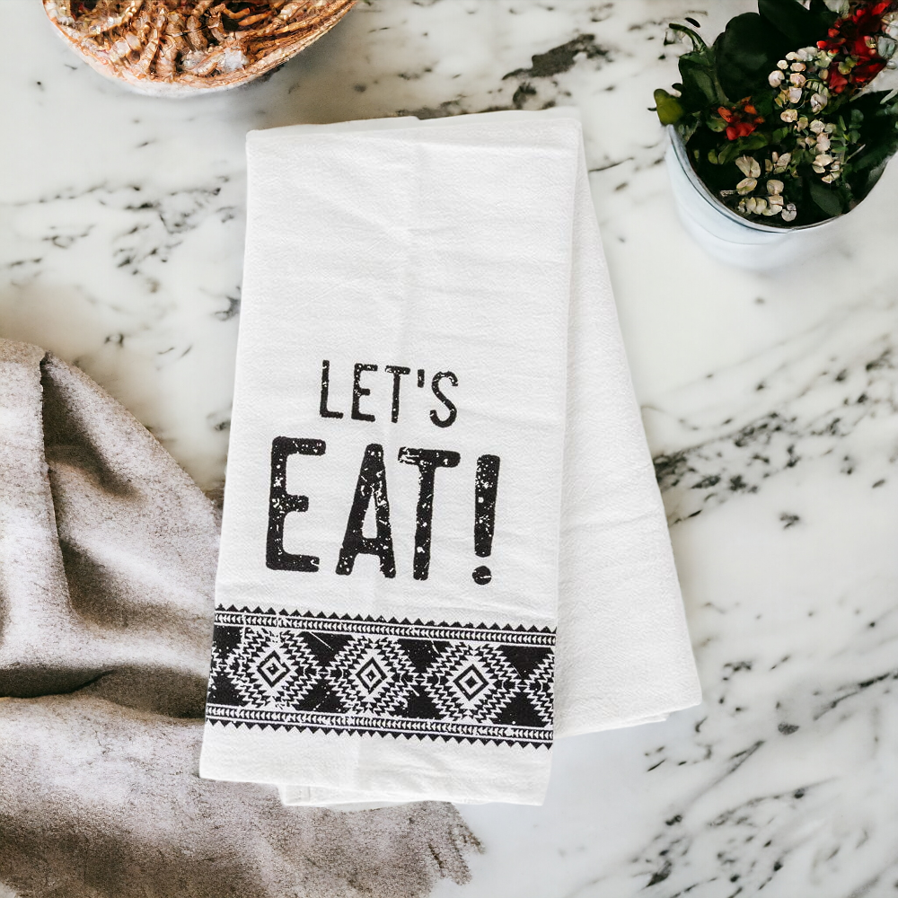 A stylish black and white tea towel featuring an Aztec design with the phrase 'Let's Eat!' printed on it, perfect for kitchen decor.