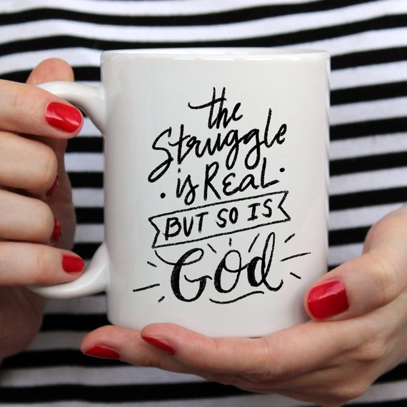 A white ceramic coffee mug featuring the phrase 'The Struggle Is Real But So Is GOD' in bold, colorful lettering, perfect for coffee lovers.