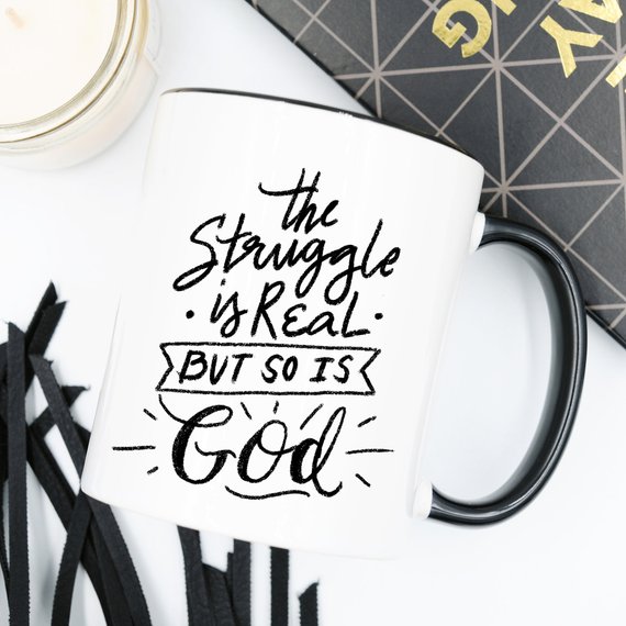 A white ceramic coffee mug featuring the phrase 'The Struggle Is Real But So Is GOD' in bold, colorful lettering, perfect for coffee lovers.