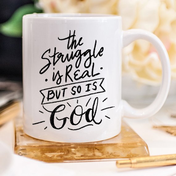 A white ceramic coffee mug featuring the phrase 'The Struggle Is Real But So Is GOD' in bold, colorful lettering, perfect for coffee lovers.