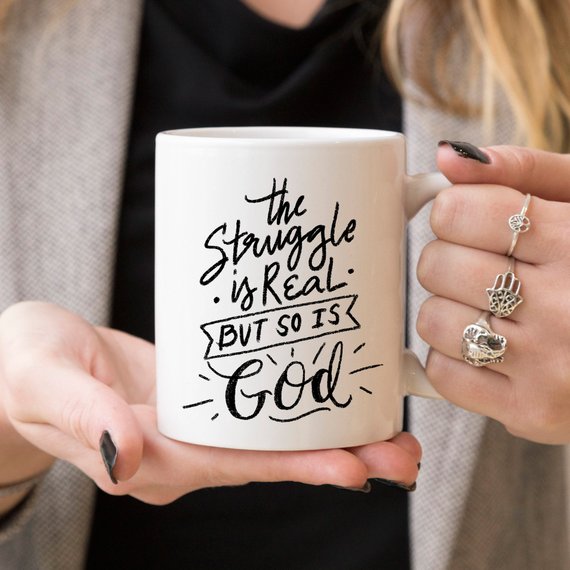 A white ceramic coffee mug featuring the phrase 'The Struggle Is Real But So Is GOD' in bold, colorful lettering, perfect for coffee lovers.