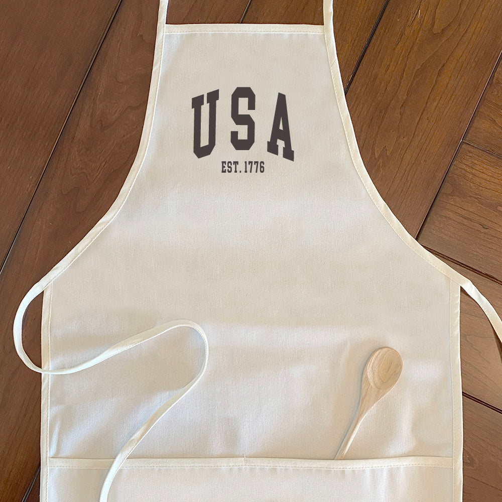 USA est 1776 Women's Apron made of cotton canvas with adjustable neck and divided front pocket, featuring elegant design and natural twill ties.