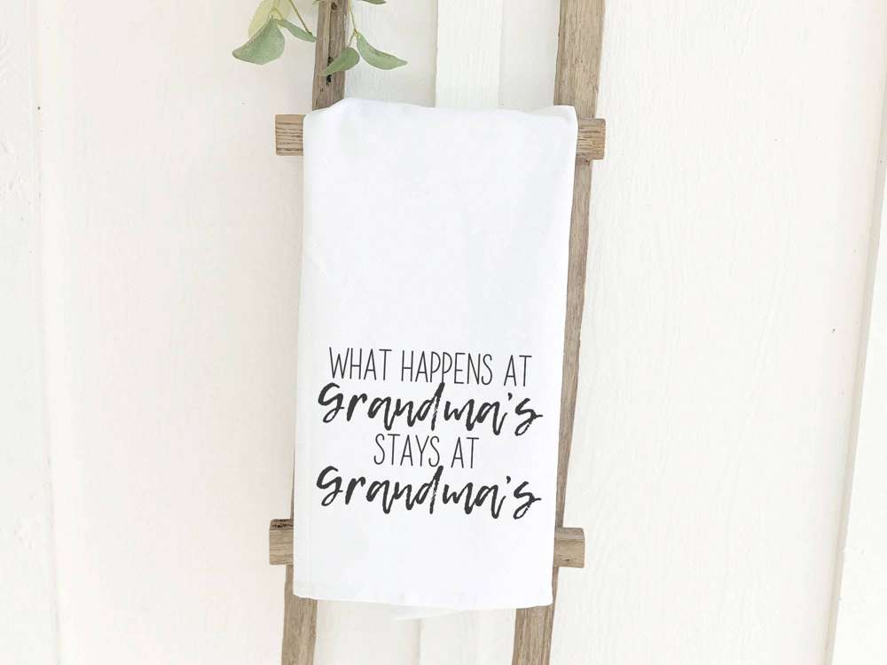 A beautifully designed cotton tea towel featuring the phrase 'What Happens at Grandma's', showcasing vibrant colors and hemmed edges.