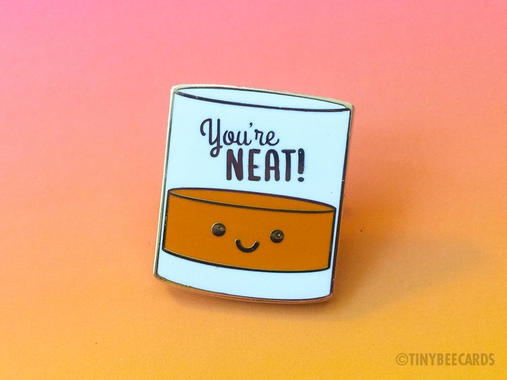 A cute enamel pin featuring a kawaii alcohol illustration with the pun text 'You're Neat!' on a gold metal background.