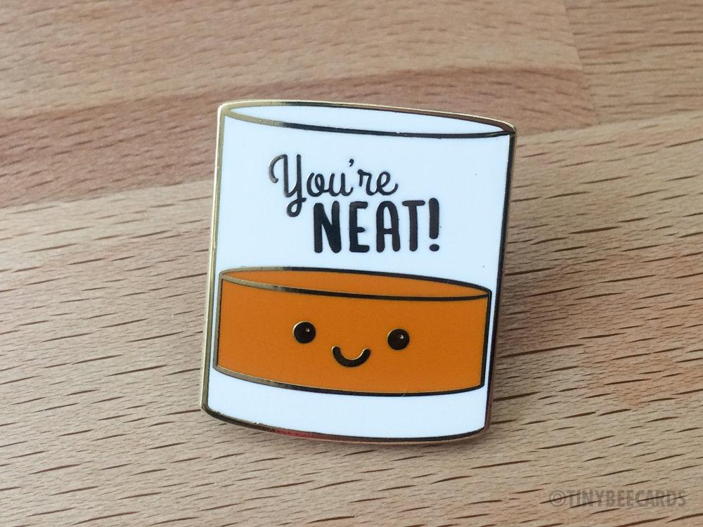 A cute enamel pin featuring a kawaii alcohol illustration with the pun text 'You're Neat!' on a gold metal background.