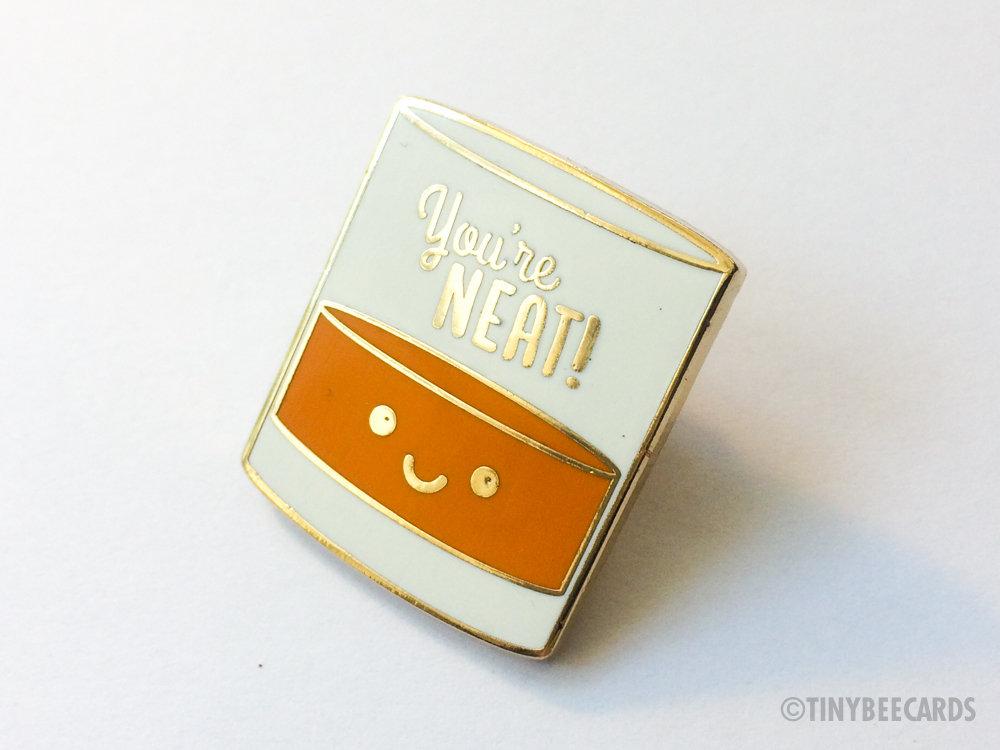 A cute enamel pin featuring a kawaii alcohol illustration with the pun text 'You're Neat!' on a gold metal background.