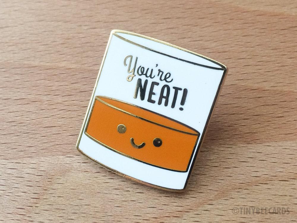 A cute enamel pin featuring a kawaii alcohol illustration with the pun text 'You're Neat!' on a gold metal background.