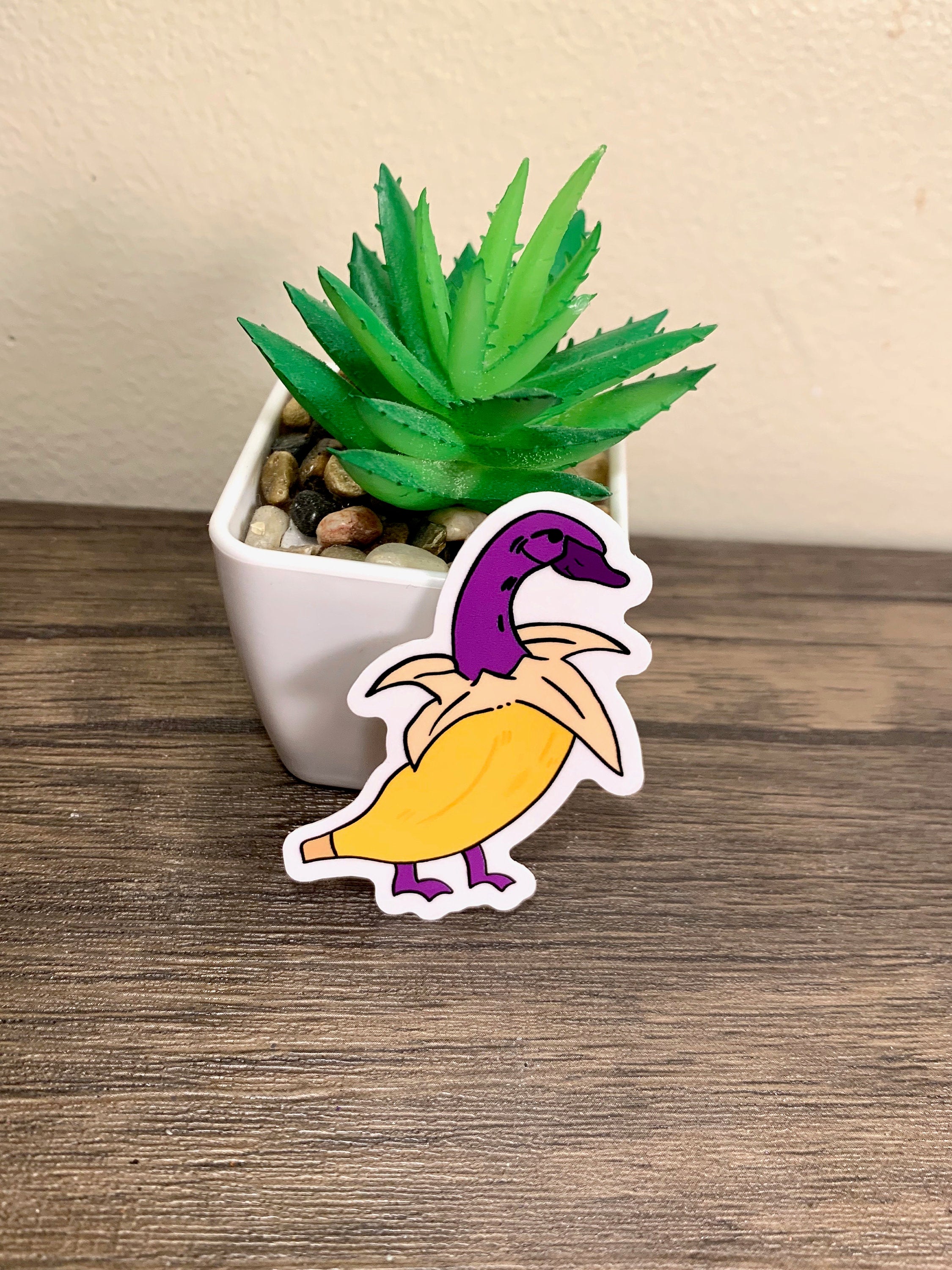 A cute banana duck sticker featuring a whimsical design, available in white and purple options, perfect for personalizing items.