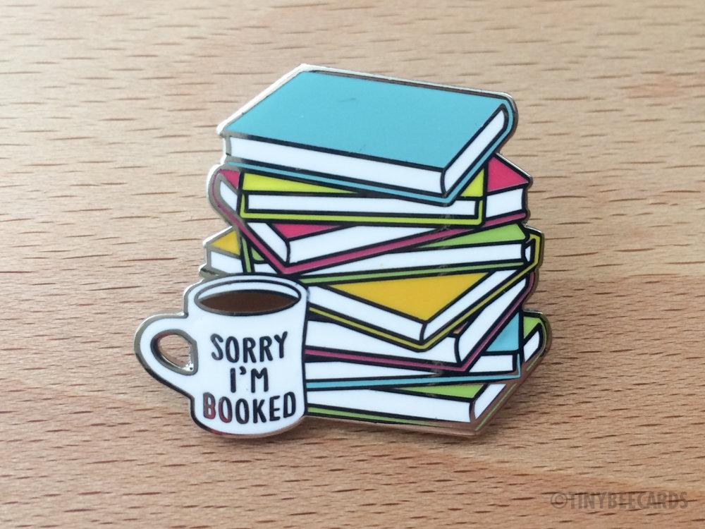 A colorful hard enamel pin featuring a stack of books and a coffee cup with the text 'Sorry I'm Booked!'