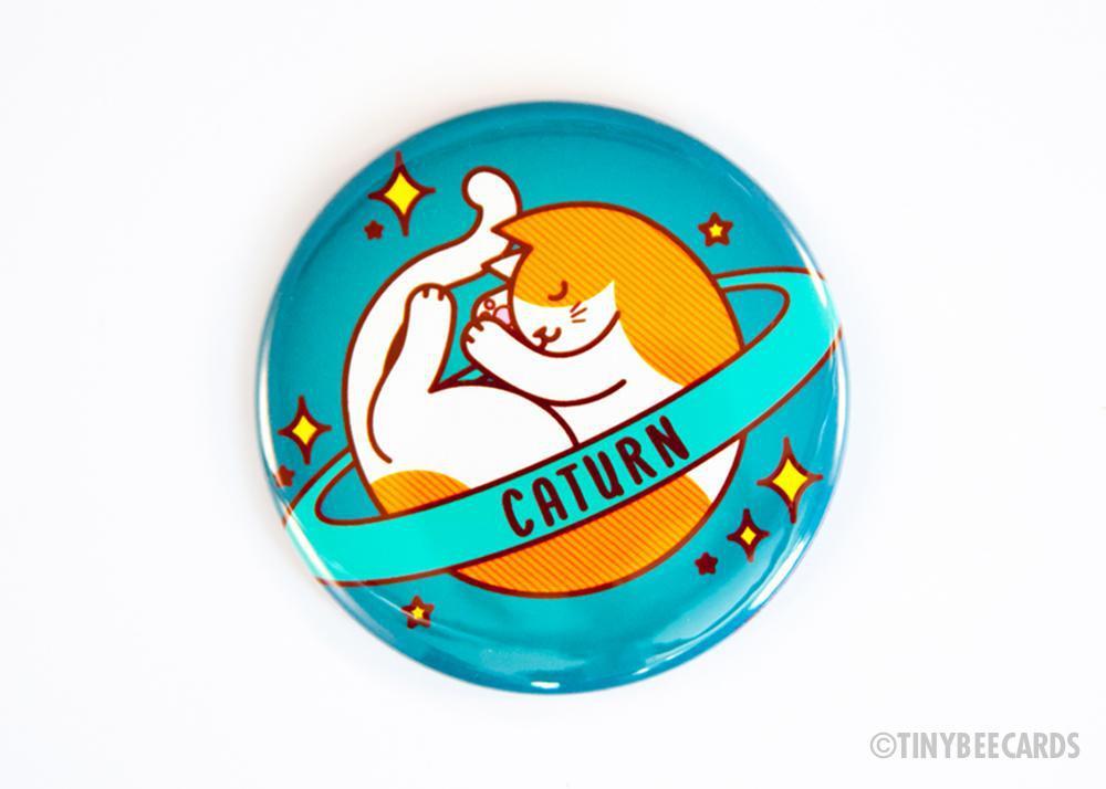 Caturn Cat Button featuring a sleepy space cat design, available as a magnet, pin, or mirror.