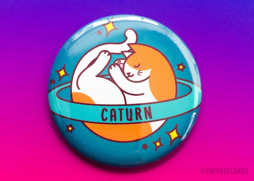 Caturn Cat Button featuring a sleepy space cat design, available as a magnet, pin, or mirror.