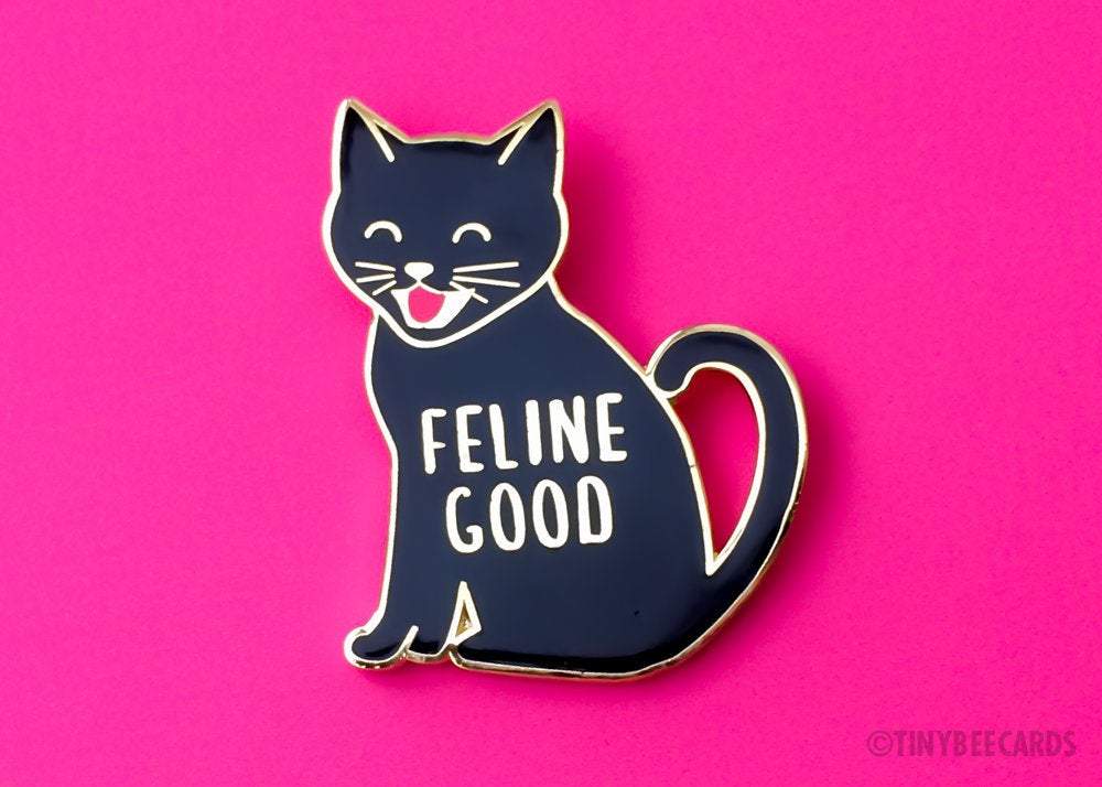 A cute enamel pin featuring a happy black cat with the text 'Feline Good' in a gold border, perfect for cat lovers.