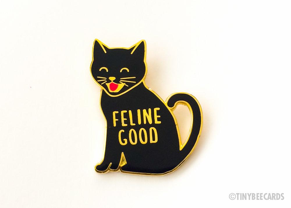 A cute enamel pin featuring a happy black cat with the text 'Feline Good' in a gold border, perfect for cat lovers.