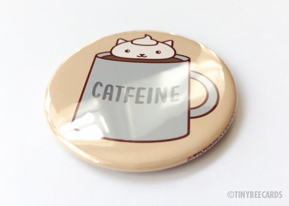 A round button featuring an adorable coffee cat design with the pun text 'Catfeine', available as a magnet or pin.