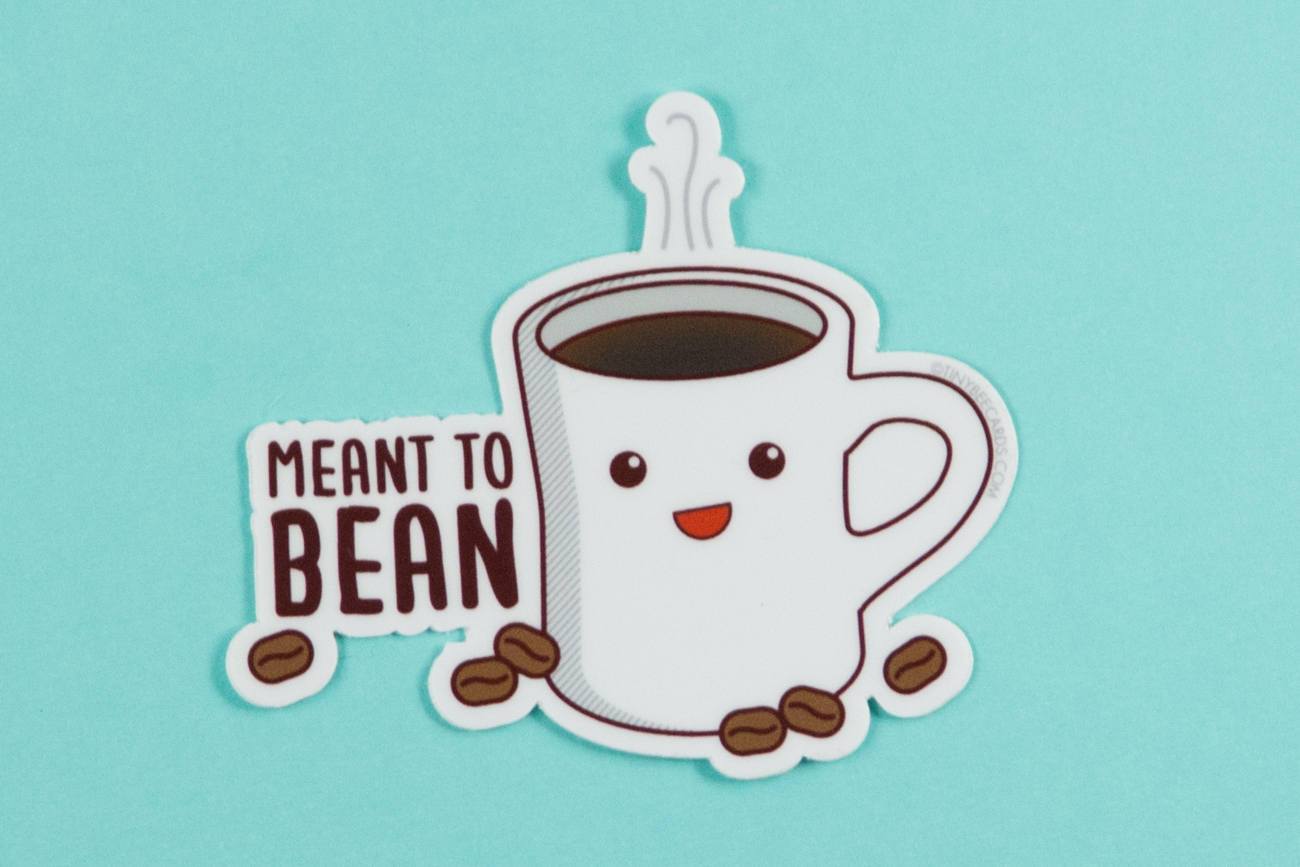 A colorful vinyl sticker featuring a happy coffee cup illustration with the pun 'Meant to Bean' in playful typography.