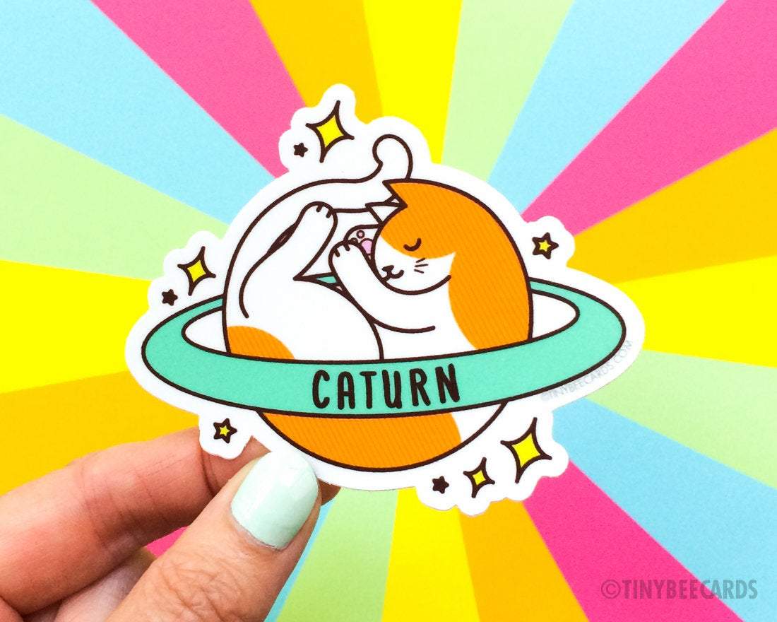 Cute Cosmic Cat Vinyl Sticker featuring an orange and white cat planet with a ring, showcasing the pun 'Caturn'.