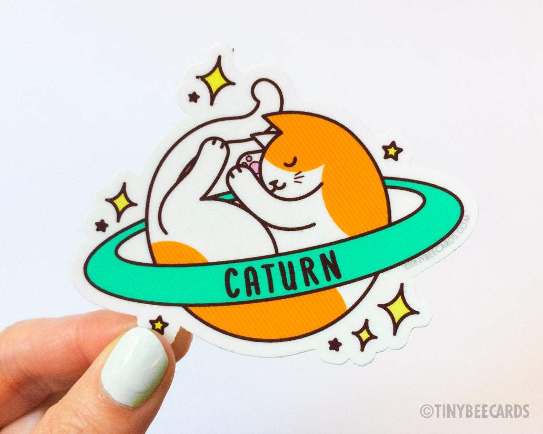 Cute Cosmic Cat Vinyl Sticker featuring an orange and white cat planet with a ring, showcasing the pun 'Caturn'.