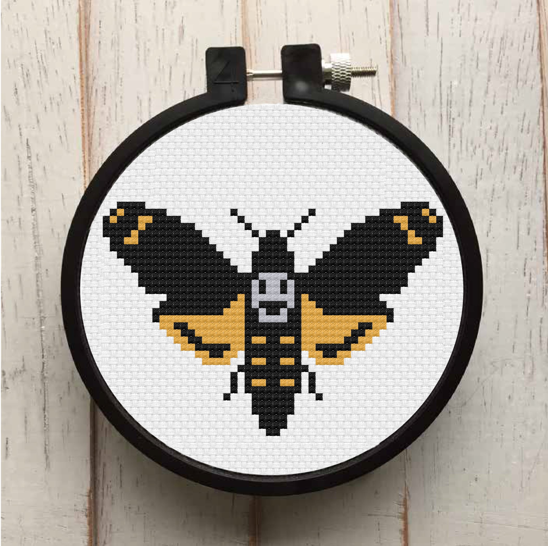 A completed counted cross stitch of a deadhead moth in black, yellow, and grey, displayed in a wooden hoop, showcasing intricate stitching details.