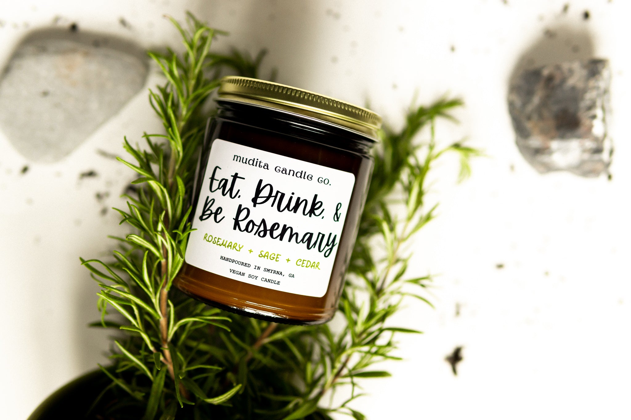 A beautifully crafted 9oz amber glass candle jar with a gold metal lid, featuring the scent 'Eat, Drink, & Be Rosemary' with notes of rosemary, sage, and cedar.