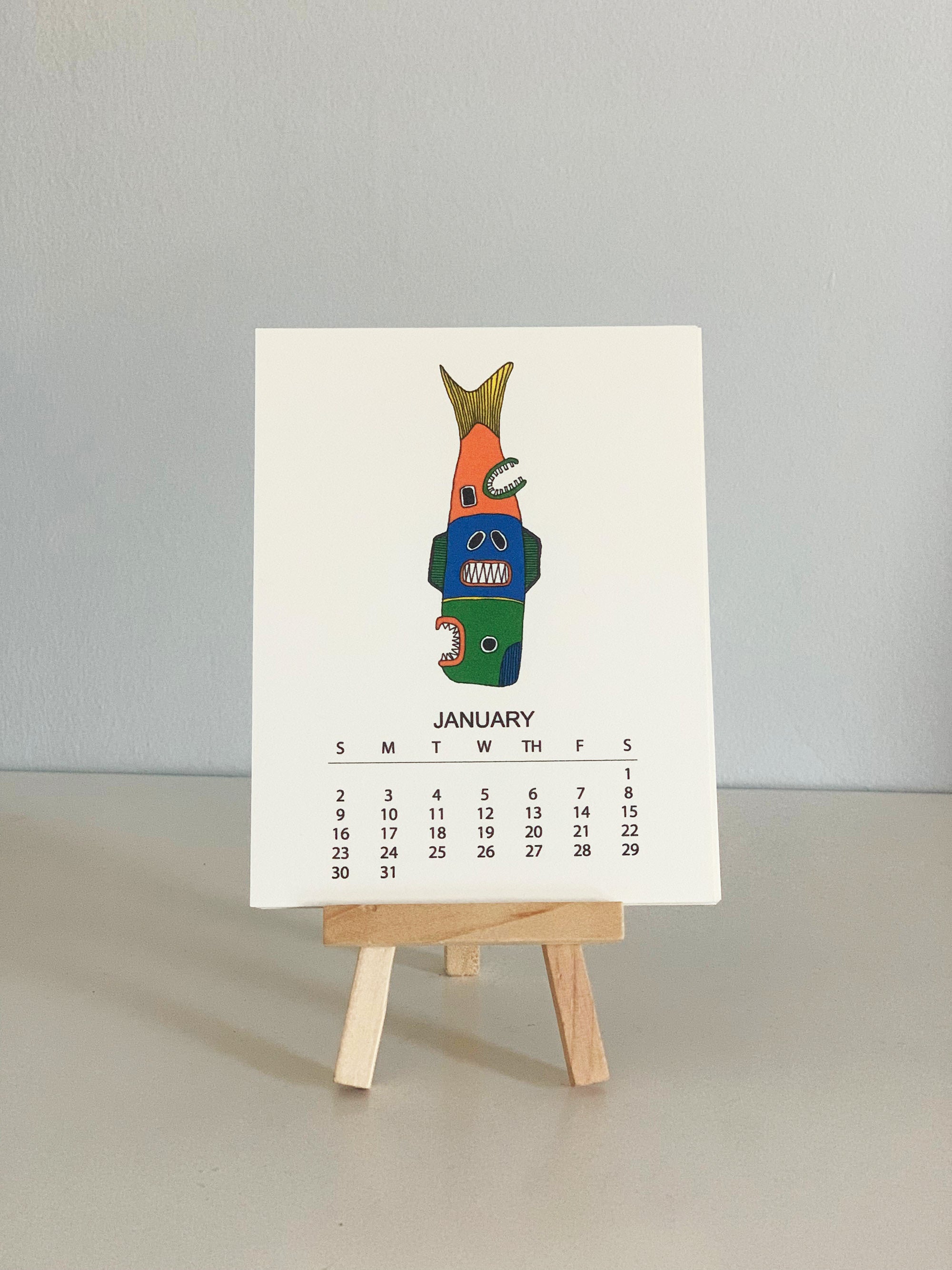 2022 Monthly Desk Calendar featuring original fish totem drawings displayed on a wooden easel.