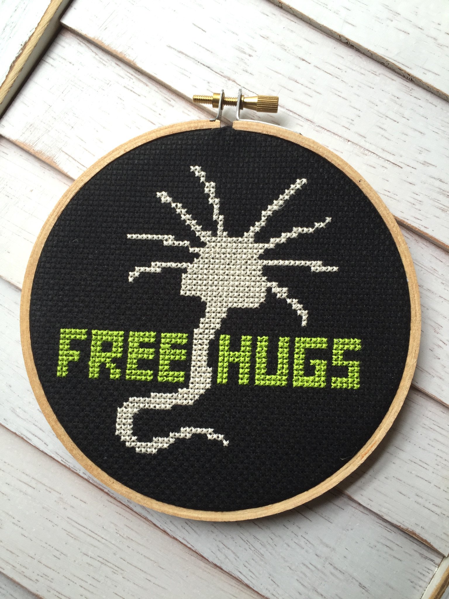 A colorful counted cross stitch kit featuring a whimsical alien character with the text 'Free Hugs', including threads, fabric, and a hoop.