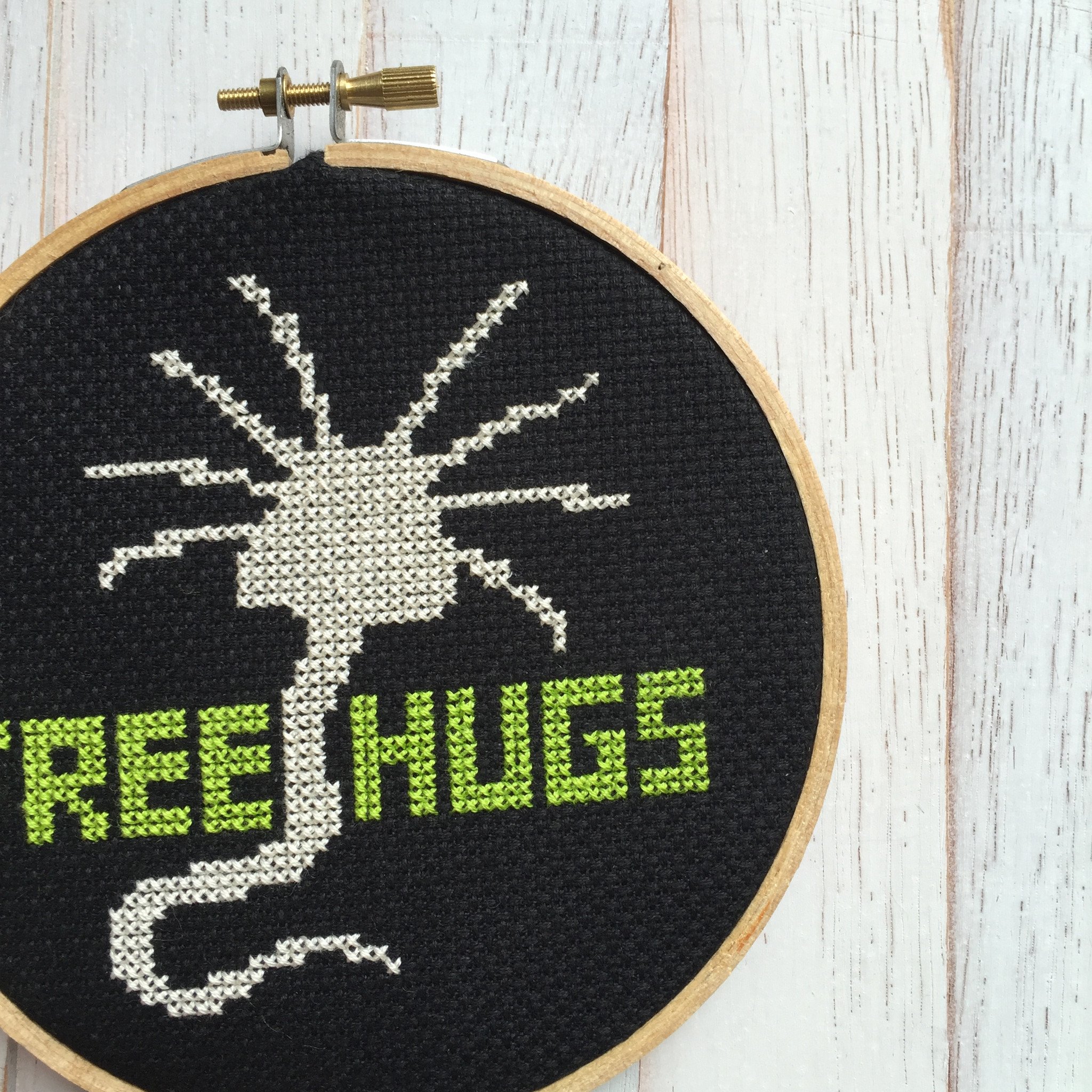 A colorful counted cross stitch kit featuring a whimsical alien character with the text 'Free Hugs', including threads, fabric, and a hoop.