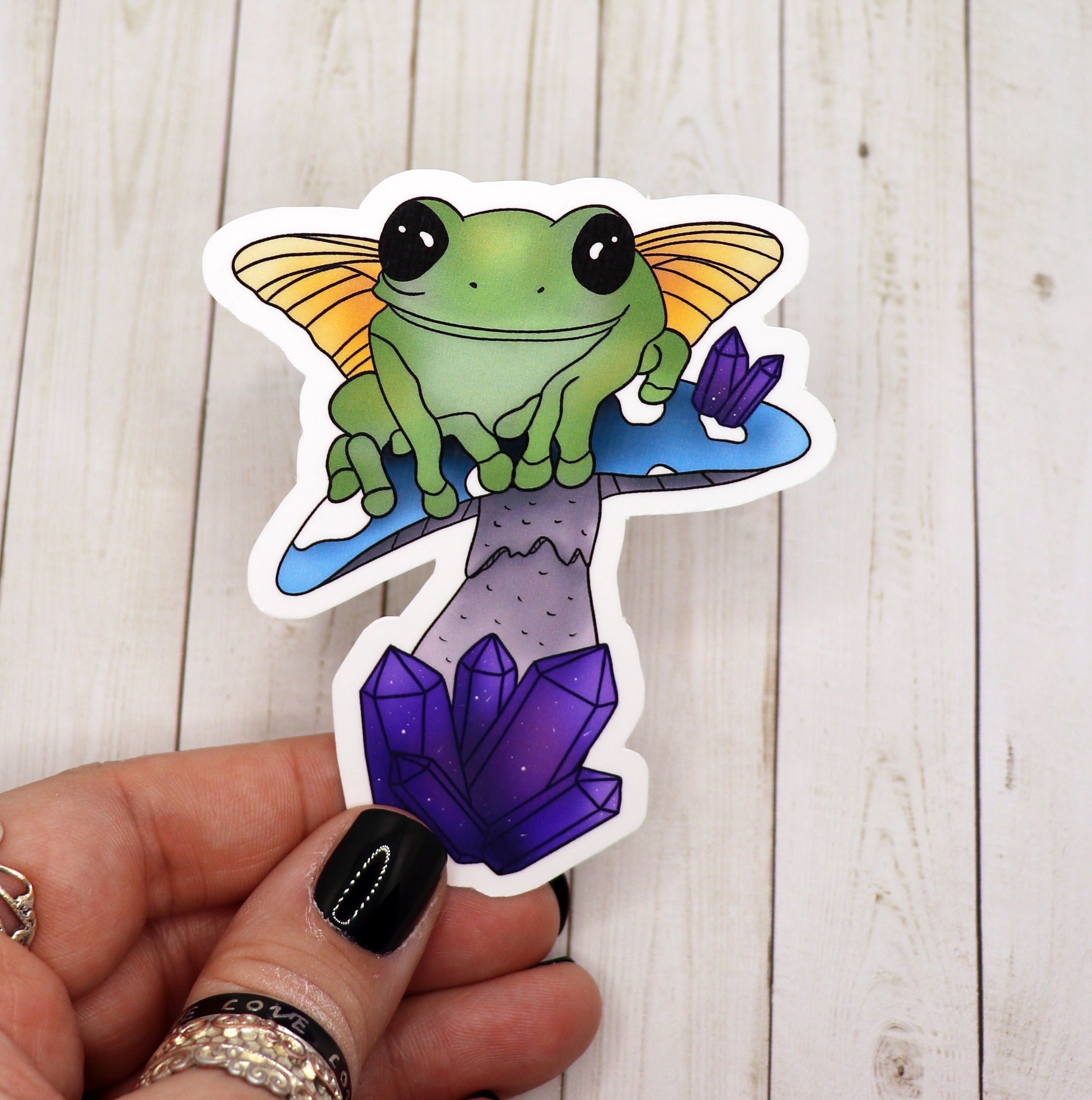 A vibrant sticker featuring a frog fairy sitting on a blue mushroom, showcasing whimsical art.