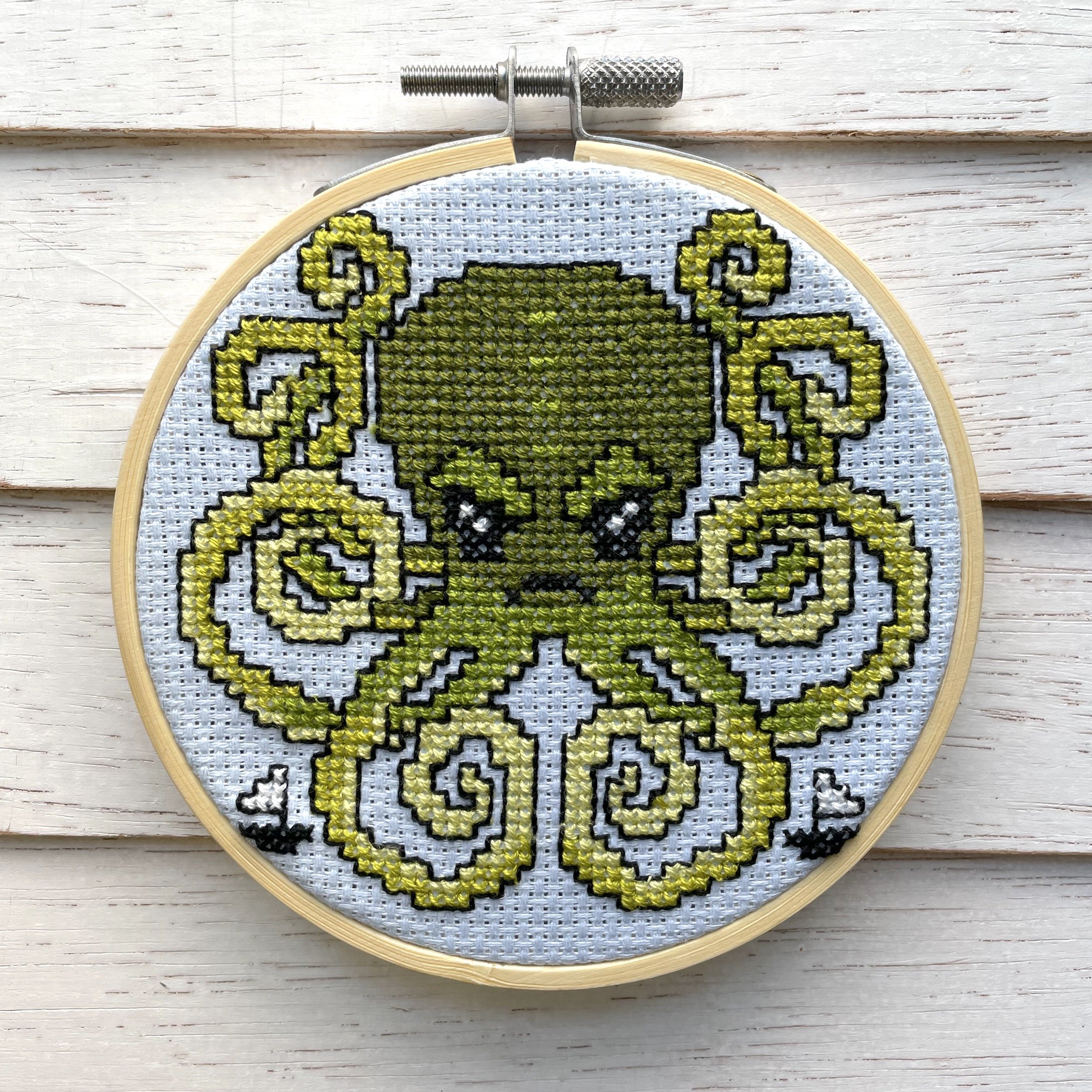 Kraken Cross Stitch Kit featuring green threads on light blue fabric, including pattern, needle, and wooden hoop.