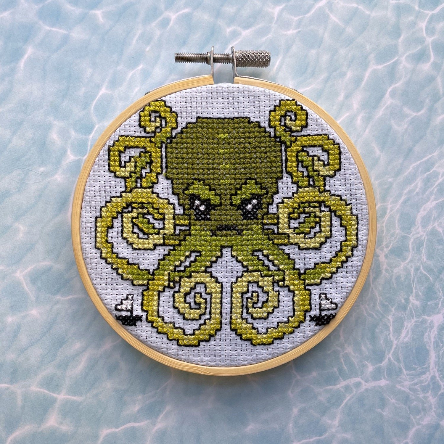 Kraken Cross Stitch Kit featuring green threads on light blue fabric, including pattern, needle, and wooden hoop.