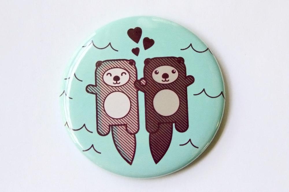 A round 2.25-inch button featuring two cute otters holding hands, designed as a refrigerator magnet or pin.