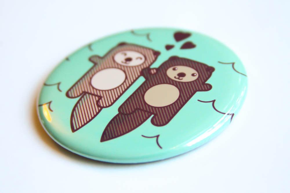 A round 2.25-inch button featuring two cute otters holding hands, designed as a refrigerator magnet or pin.