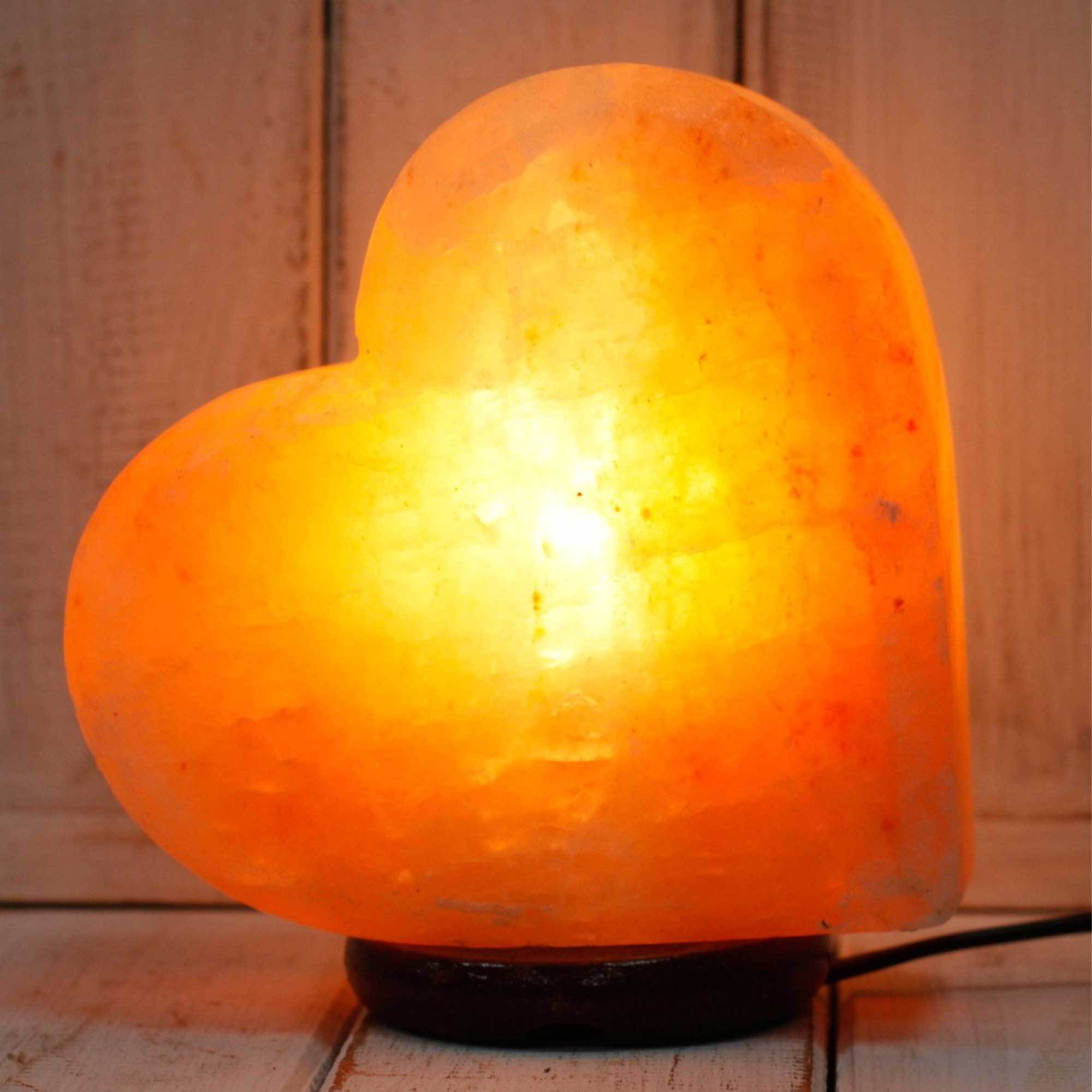 A beautifully handcrafted heart-shaped Himalayan pink salt lamp with a warm glow, resting on a wooden base, showcasing natural variations in color.