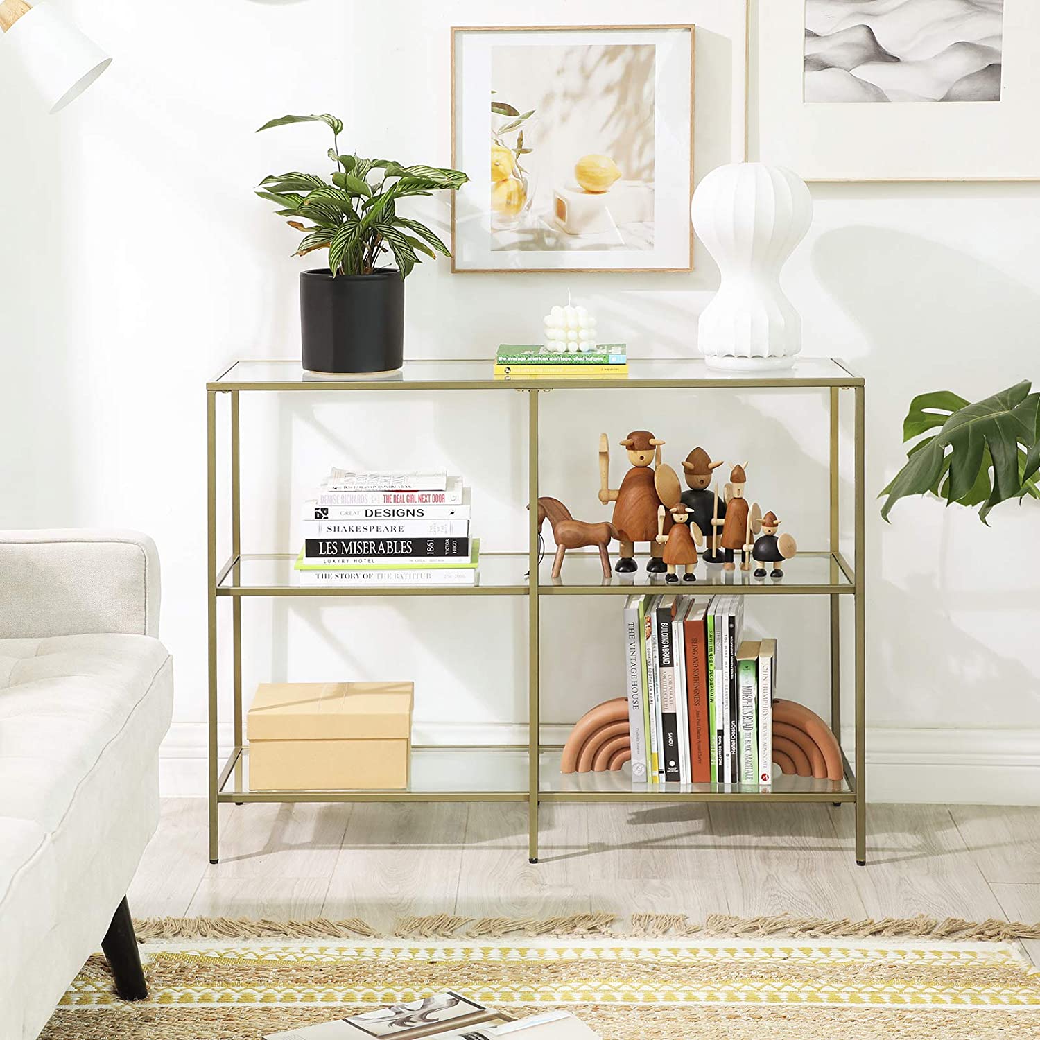 A modern 3-tier tempered glass sofa table with a sleek golden metal frame, showcasing books and decorative plants.