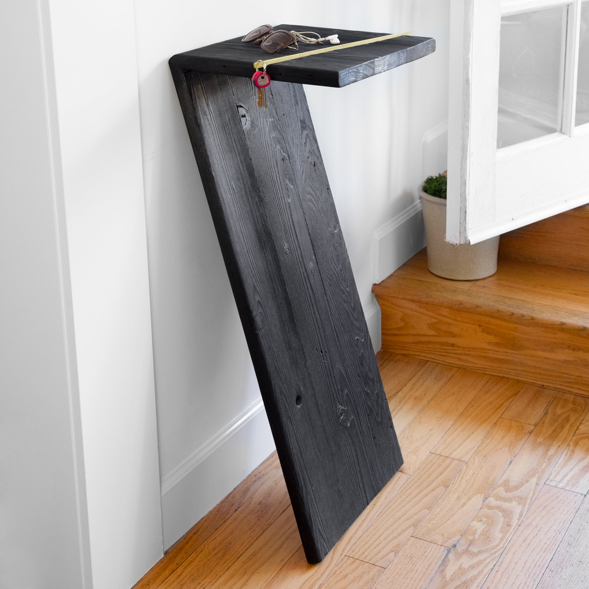 The 7 console table featuring a charred wood finish and brass accent, leaning against a wall, showcasing its unique design.