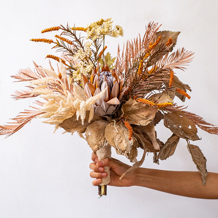 A beautifully arranged Artificial Silk Natural Fall Designer Flower Bouquet featuring protea and fern plants in white, purple, and light brown colors.